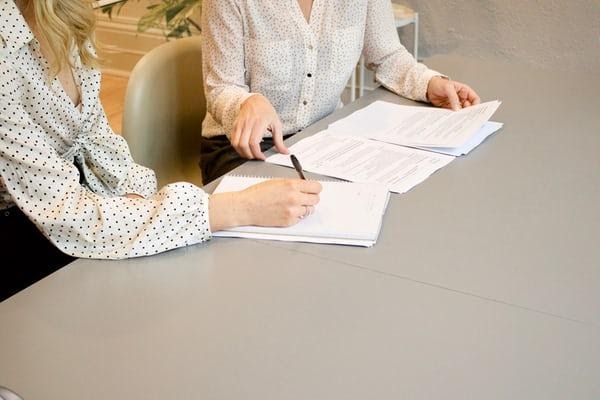 Two women sat at a table, one with a pen in hand about to sign a document.