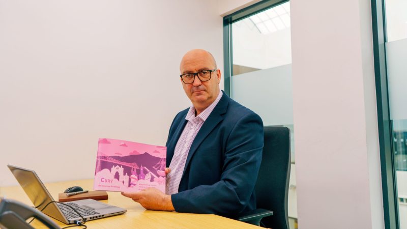Head of CURV John Holden sat at a desk, holding a copy of the new CURV response strategy.