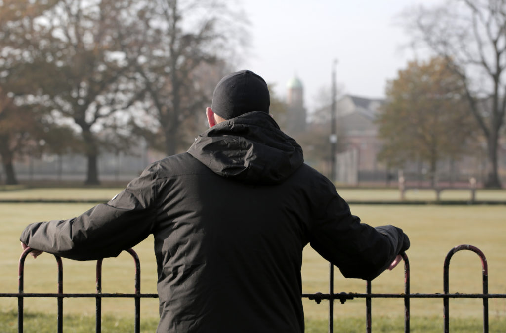 A man in a black coat and black hat with his back to the camera looking across a field.