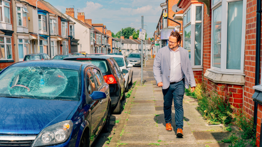 PCC Matt Storey walking on a pavement down a street. He is turning to look at a blue car with a smashed windscreen.