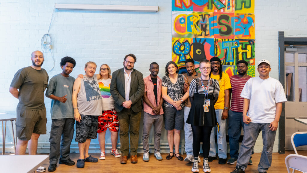 Police and Crime Commissioner Matt Storey is stood with people at the Hart Gables office. They are stood either side of the PCC in front of a blue wall with a mural on it. They are smiling.