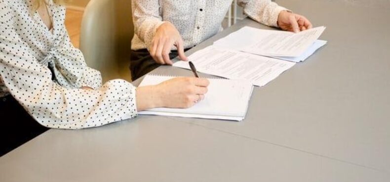 Two women sat at a table, one with a pen in hand about to sign a document.