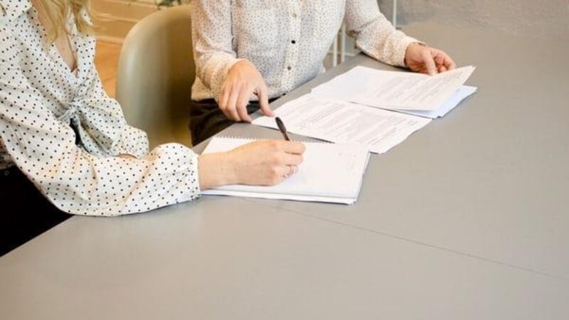 Two women sat at a table, one with a pen in hand about to sign a document.