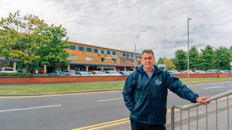 PCC Steve Turner stood in front of a parade of shops in Middlesbrough. He is wearing a blue fleece and has one hand on a metal railing.
