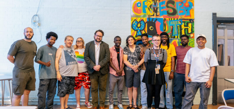 Police and Crime Commissioner Matt Storey is stood with people at the Hart Gables office. They are stood either side of the PCC in front of a blue wall with a mural on it. They are smiling.