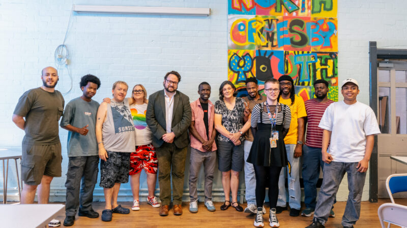 Police and Crime Commissioner Matt Storey is stood with people at the Hart Gables office. They are stood either side of the PCC in front of a blue wall with a mural on it. They are smiling.