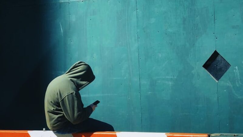 A person sat on a railing with their hood up. They are slumped down looking at their mobile phone.