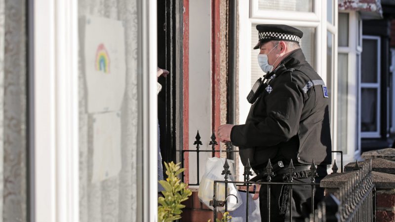 A police officer in uniform stands on a doorstep speaking to an unseen resident